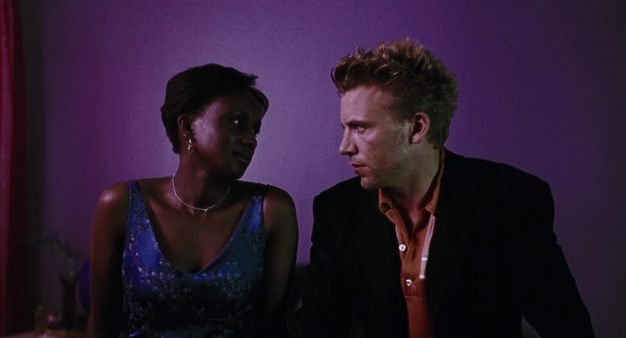 Last Night (1998) movie still: medium — A man and woman sit together in front of a solid, deep purple wall. They turn their heads to maintain eye…