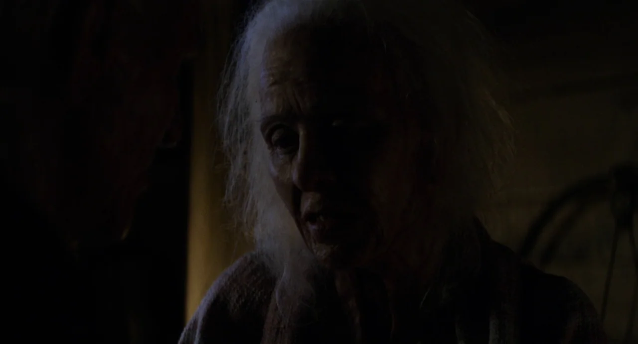 X (2022) movie still: closeup — An elderly person is lit by a dim light source inside a dark space. The subject stares toward another person who is…