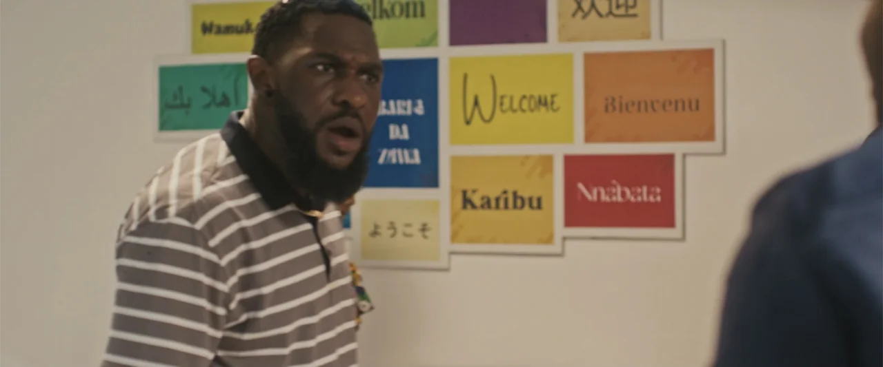Casa De Novia (2024) movie still: medium — An interior office space features a white wall covered with multicolored welcome signs. A man in a…