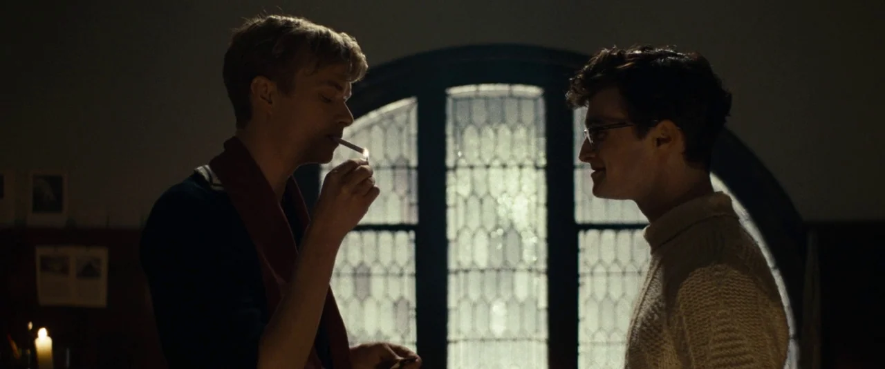 Kill Your Darlings (2013) movie still: medium — A dark indoor setting features a large circular leaded glass window in the background. Two men stand…