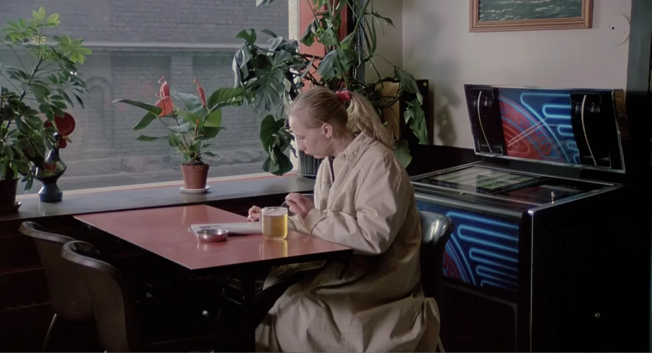 The Match Factory Girl (1990) movie still: medium — A booth in a quiet diner features a red table, large plants, and a jukebox. A woman sits in a…