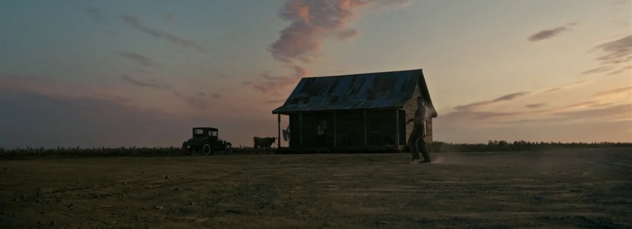 Sinners (2025) movie still: wide — A remote rural homestead sits on a flat, dusty plain during golden hour. A shirtless man walks through the dirt…