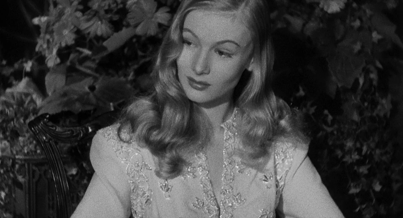 Dead Men Don’t Wear Plaid (1982) movie still: medium — A woman sits before a dark, dense mass of thick leafy plants and flowers. She looks…