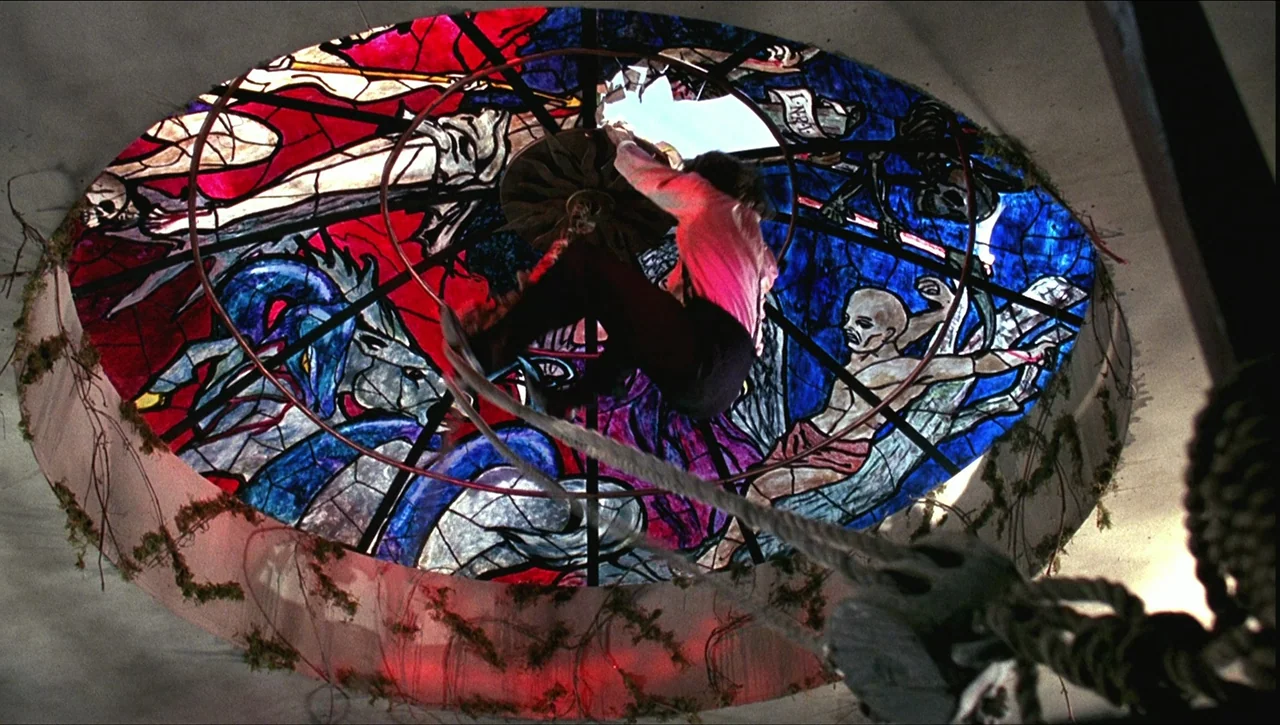 Necronomicon: Book of Dead (1993) movie still: medium — A man hangs from a central support inside a large circular stained glass dome. The stained…