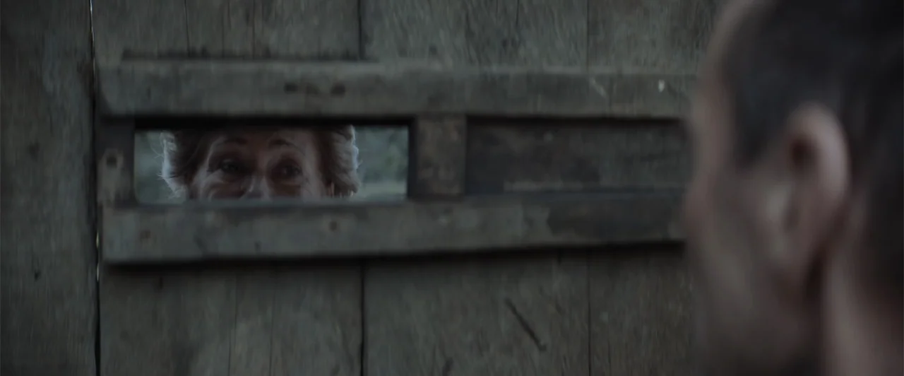 My Neighbor Adolf (2022) movie still: medium — A weathered wooden gate with a horizontal viewing slat sits outdoors. An elderly woman is visible…