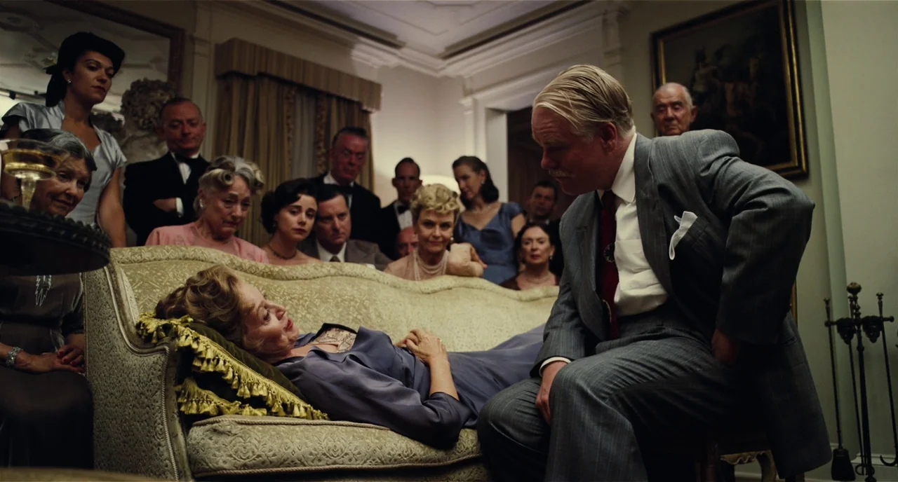 The Master (2012) movie still: medium — An elegant 1950s interior space filled with guests gathered around a central beige sofa. A man sits by a…