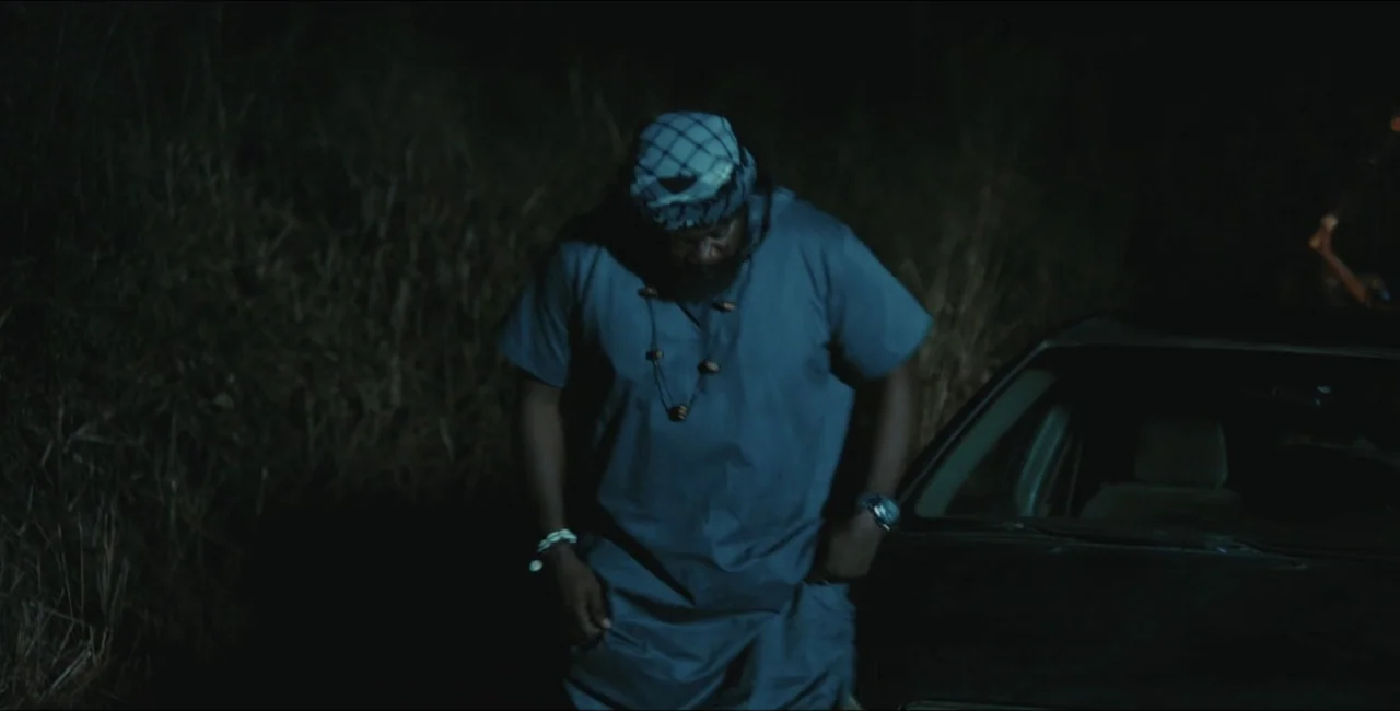 Project Assault (2025) movie still: medium — A dark outdoor space at night features dry tall grass and a vehicle. A man wearing a head covering…