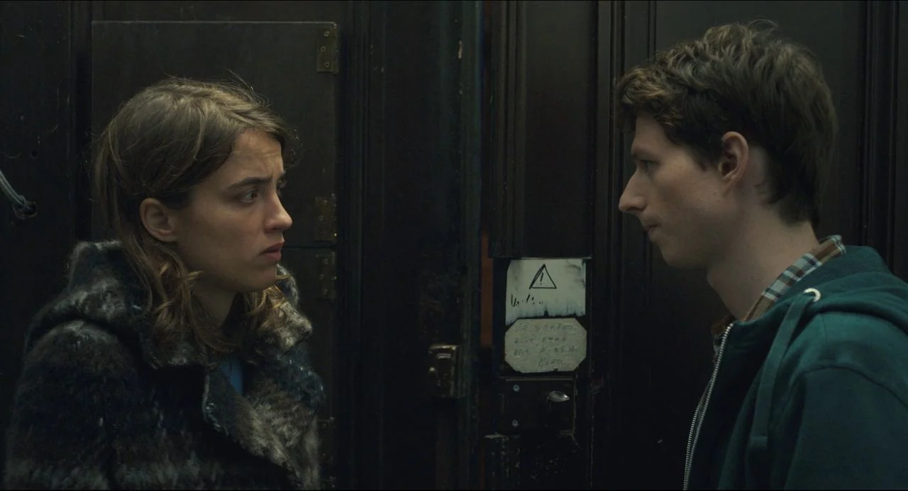 The Unknown Girl (2016) movie still: medium — Two individuals stand facing each other before a dark paneled door with metal hinges. A small…