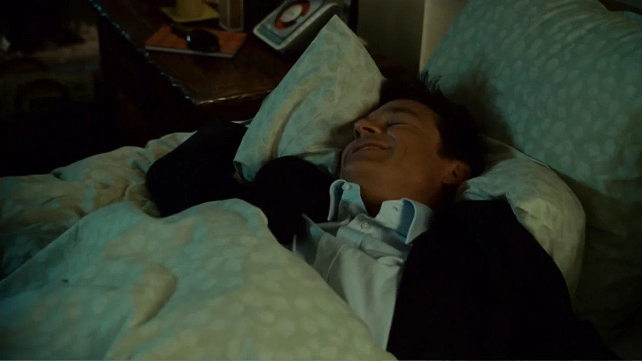 Hancock (2008) movie still: closeup — A dimly lit bedroom with a patterned bedspread and a wooden nightstand. A man in a dress shirt and blazer…