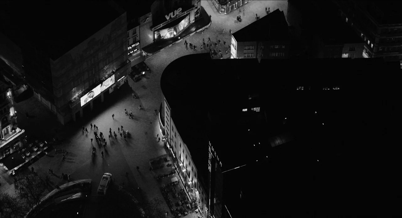 One More Time With Feeling movie still: establishing — An aerial view of a city square at night with several illuminated building facades. Small…