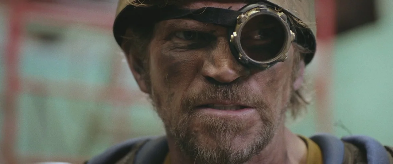 Turbo Kid (2015) movie still: closeup — A close-up of a weathered man in a cap and single goggle lens. The man stares forward with a furrowed brow…