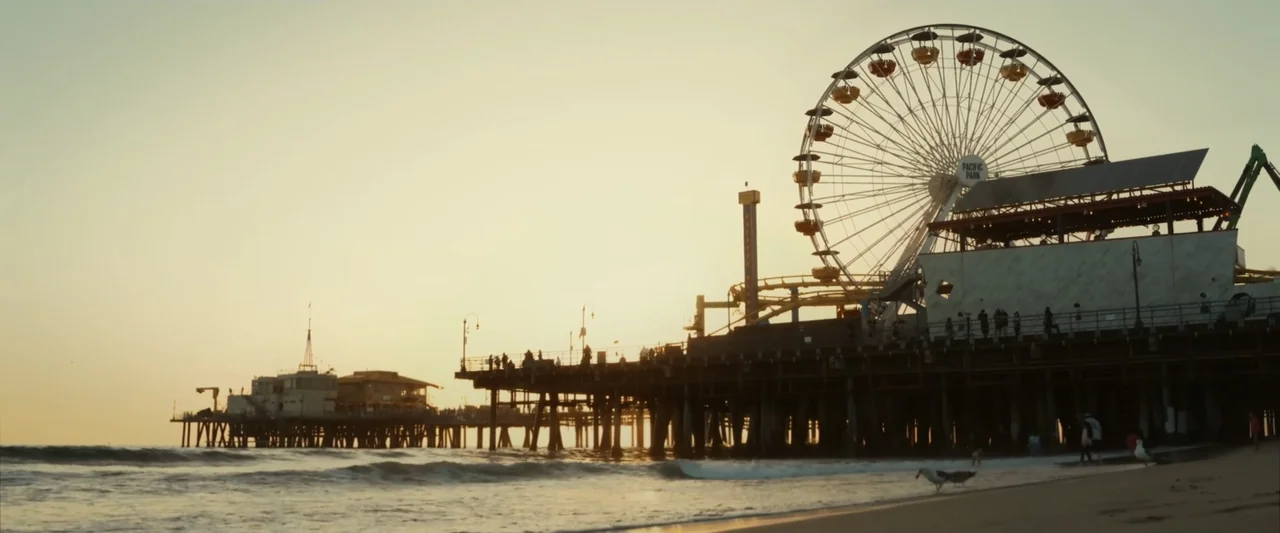 I Love America (2022) movie still: establishing — A wooden amusement pier extends over the ocean at sunset with a large ferris wheel. People walk…