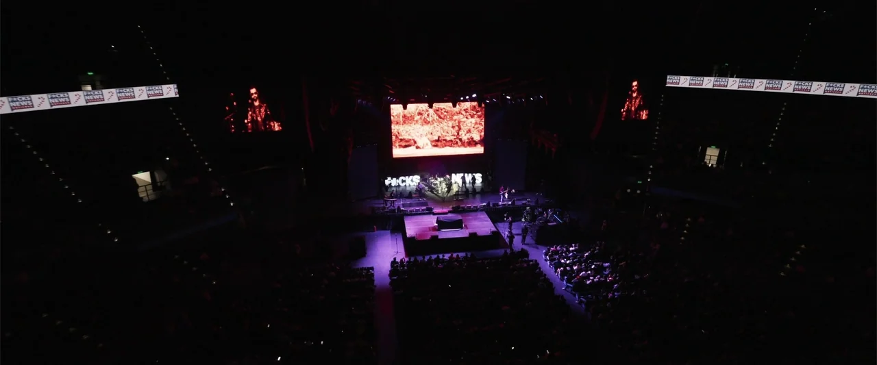 Making of: F*CKS NEWS (2025) movie still: wide — A high-angle view of a large, dark concert arena during a live musical performance. The stage…