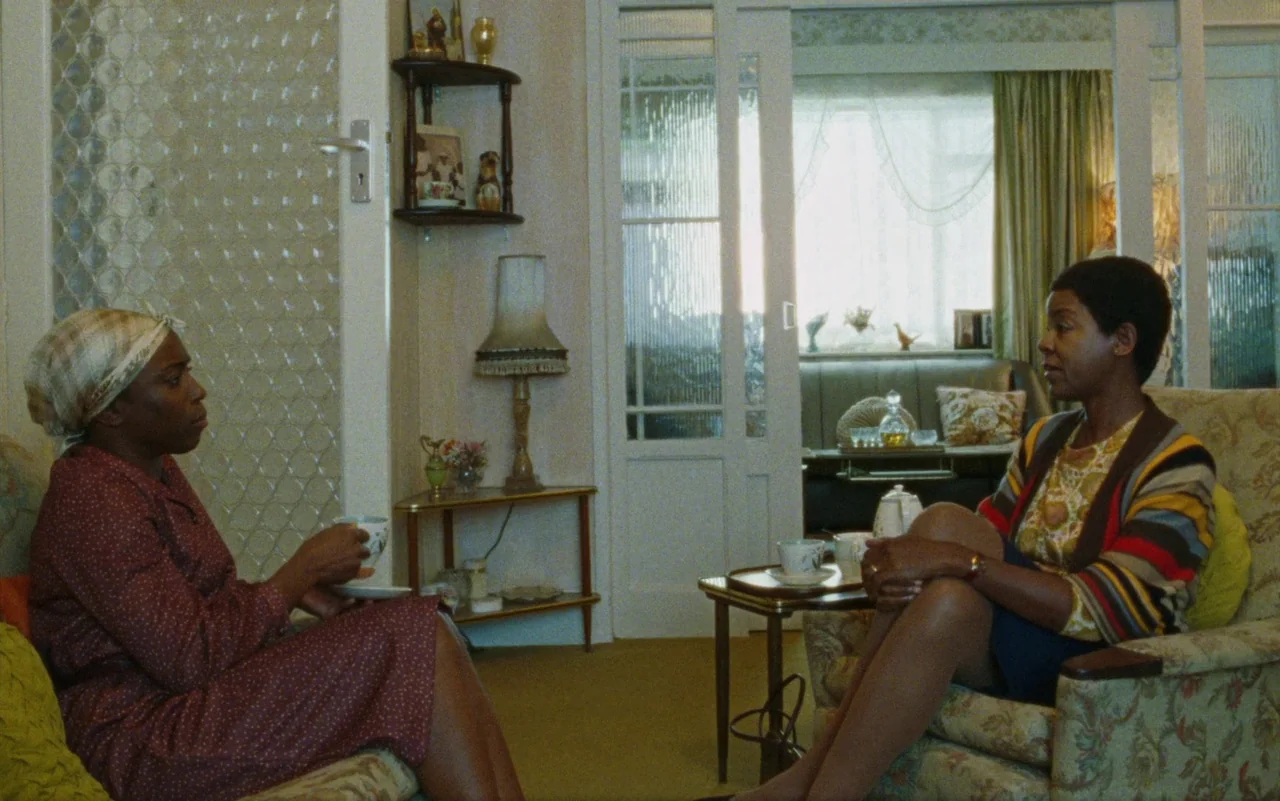 Education (2020) movie still: medium — A 1980s domestic interior features patterned wallpaper, floral upholstery, and a glass-paneled room divider.…
