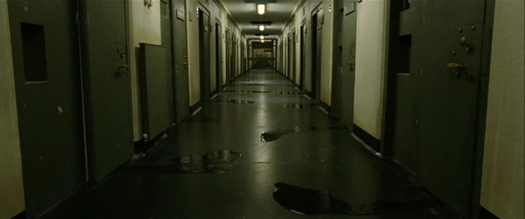 Hunger (2008) movie still: establishing — A long, symmetrical prison corridor is lined with uniform metal doors on both sides. Dark liquid puddles…