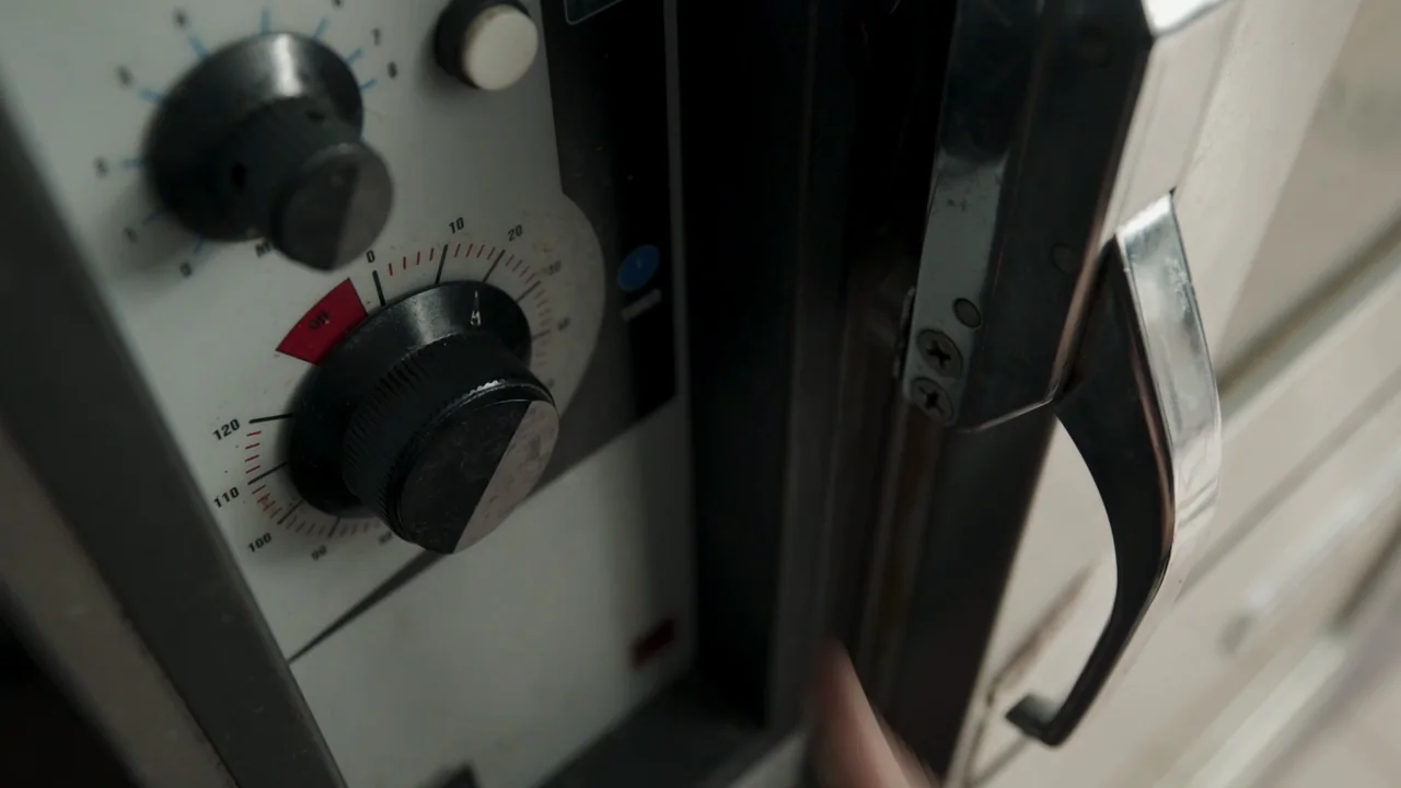A Gingerbread Christmas (2022) movie still: closeup — A close-up view shows the control dial and metal handle of a commercial oven. A blurred hand…