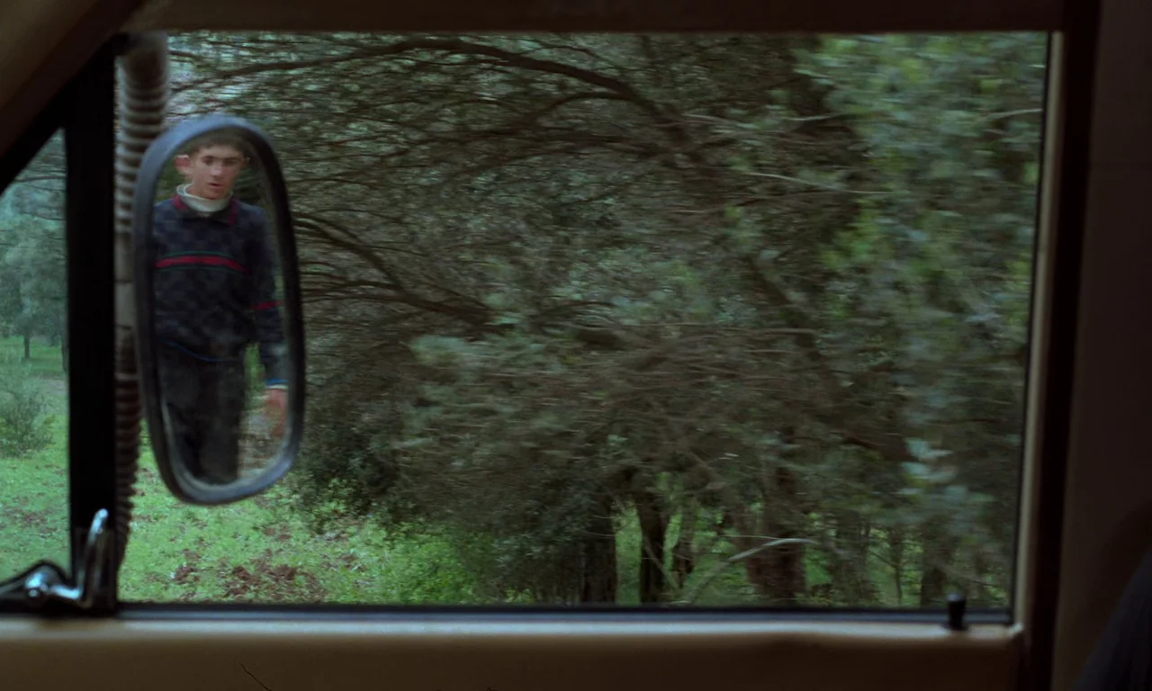 Through The Olive Trees (1994) movie still: wide — A view through a car passenger side window showing a wooded area outside. A young boy walks along…