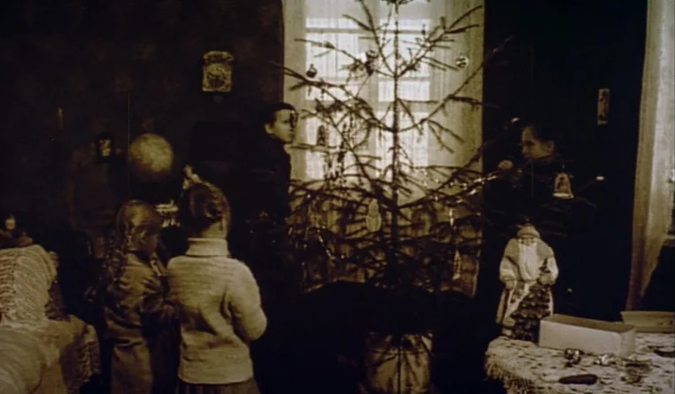 The Turn of the Century (2001) movie still: wide — A dimly lit room holds a sparse Christmas tree positioned before a bright window. Several…