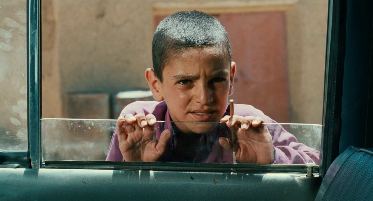 The Wind Will Carry Us (1999) movie still: closeup — An exterior view of a young boy peering through the window of a vehicle. He rests his hands on…
