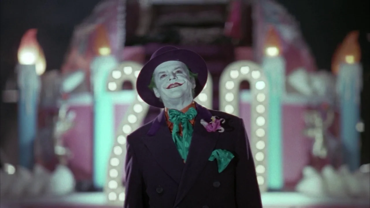 Batman (1989) movie still: medium — A man in theatrical makeup stands before a large, brightly lit parade float. He looks toward the side while…