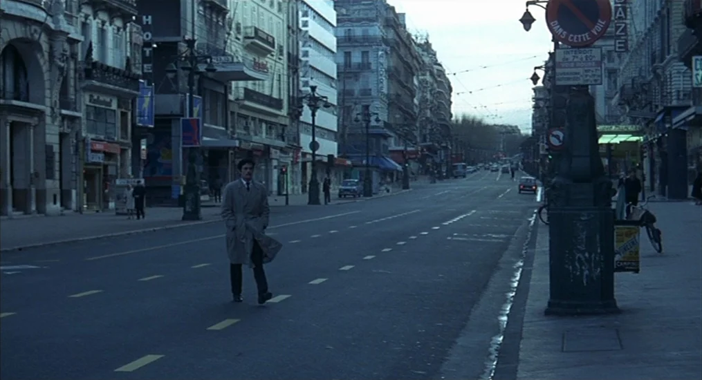 Le Cercle Rouge (1970) movie still: wide — A wide city street lined with European apartment buildings and commercial signage at dawn. A lone man in…
