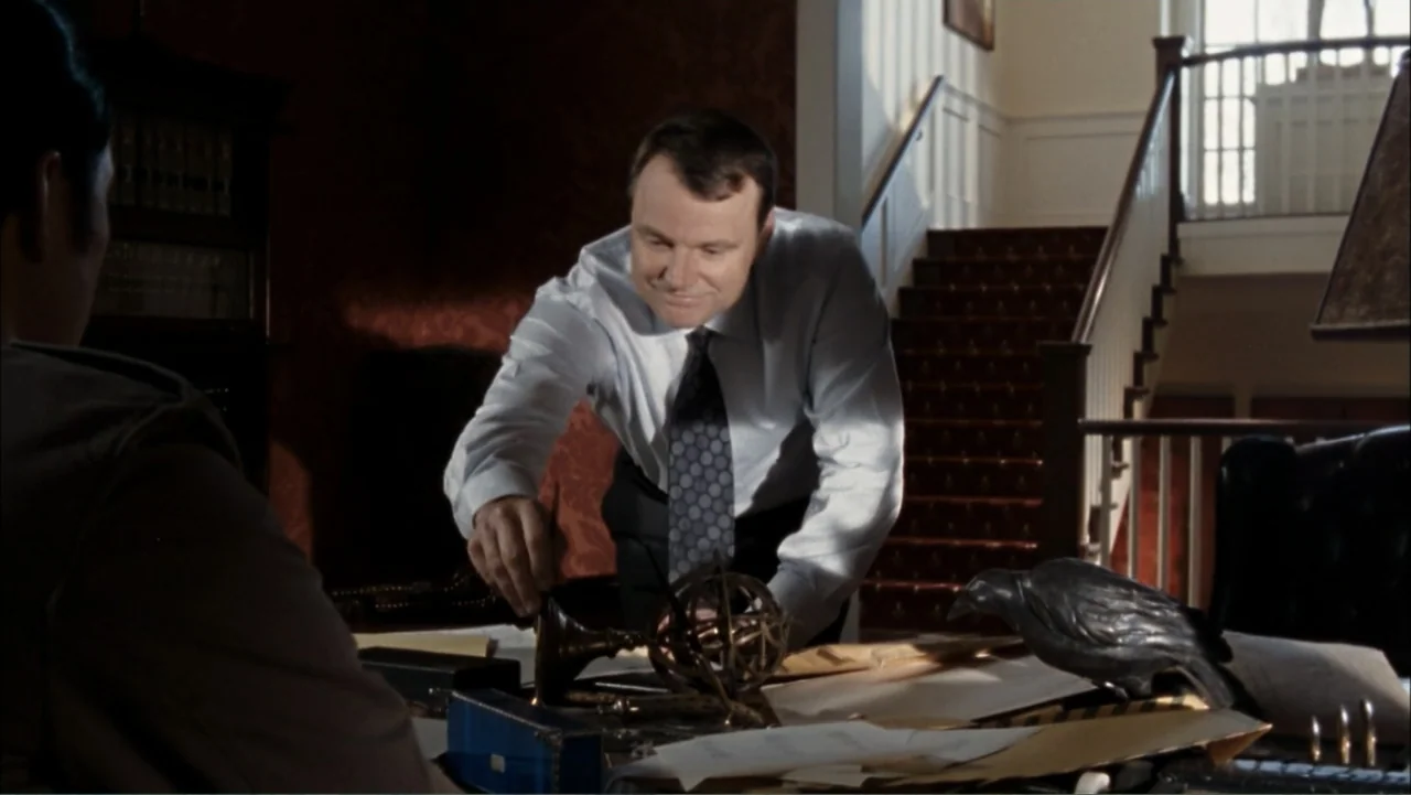 Driven to Kill (2009) movie still: medium — A well-appointed home study features red wallpaper and a wooden staircase in the background. A man in a…