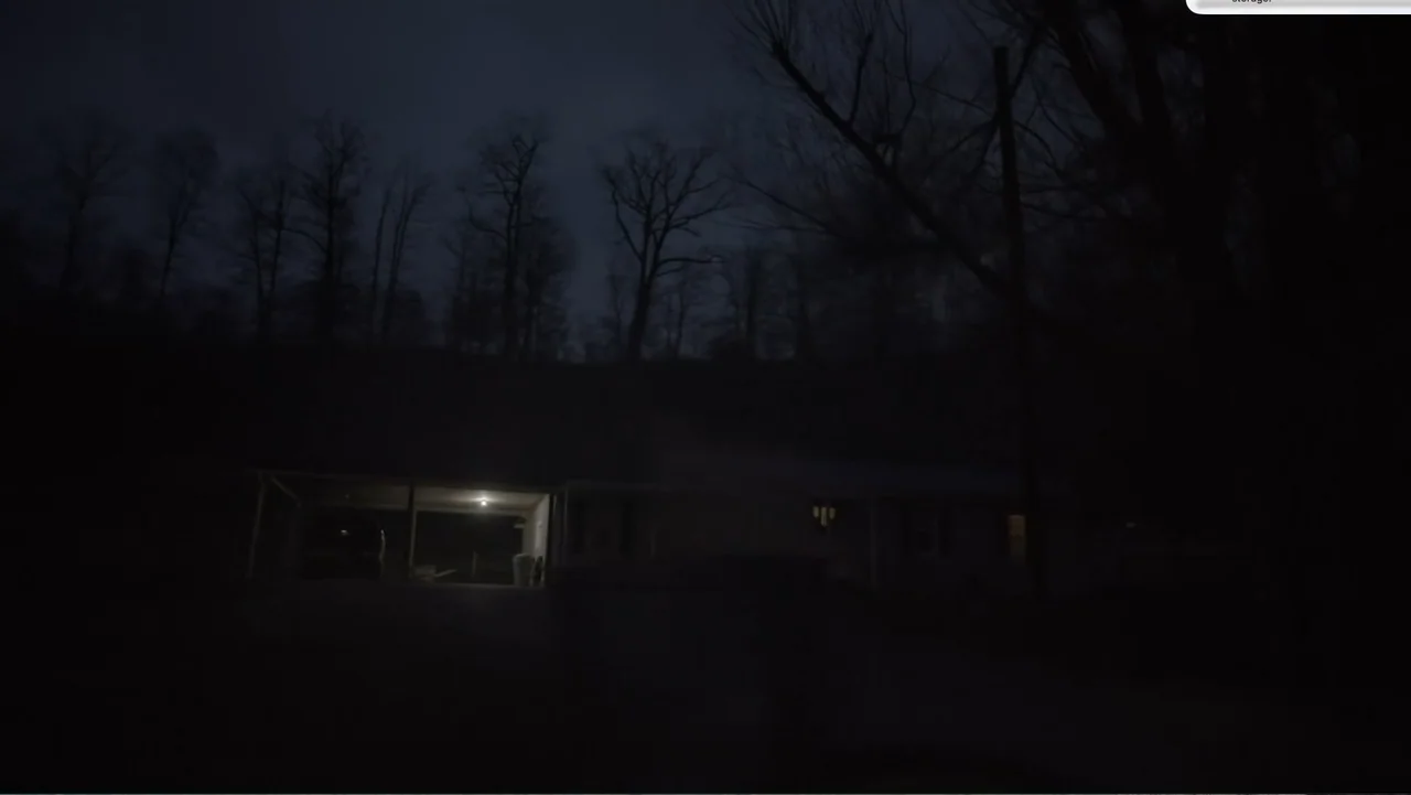 Murder at Carter County Mines (2023) movie still: wide — A suburban house sits at the base of a dark, wooded hill at night. A bright porch light…