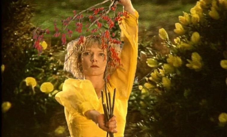 The Garden (1990) movie still: medium — A woman stands in a lush green space filled with yellow flowering shrubs. She holds a thin branch with small…