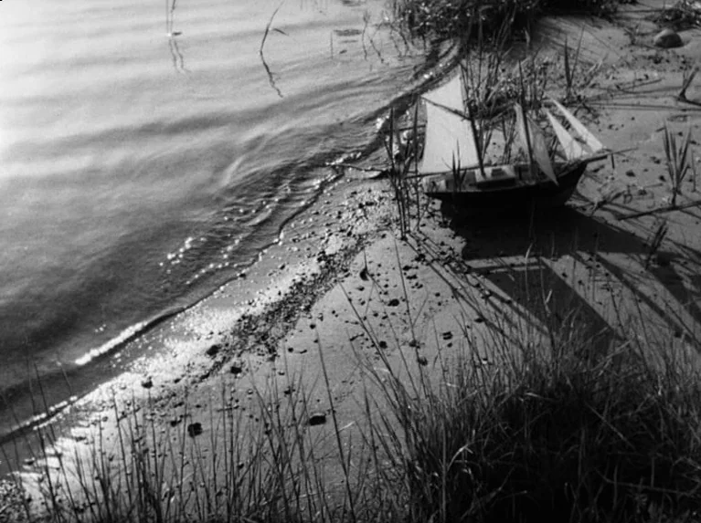 Waiting Women (1952) movie still: wide — A small toy sailboat sits on a sandy, grassy shore by the water. Gentle waves lap at the beach as sunlight…
