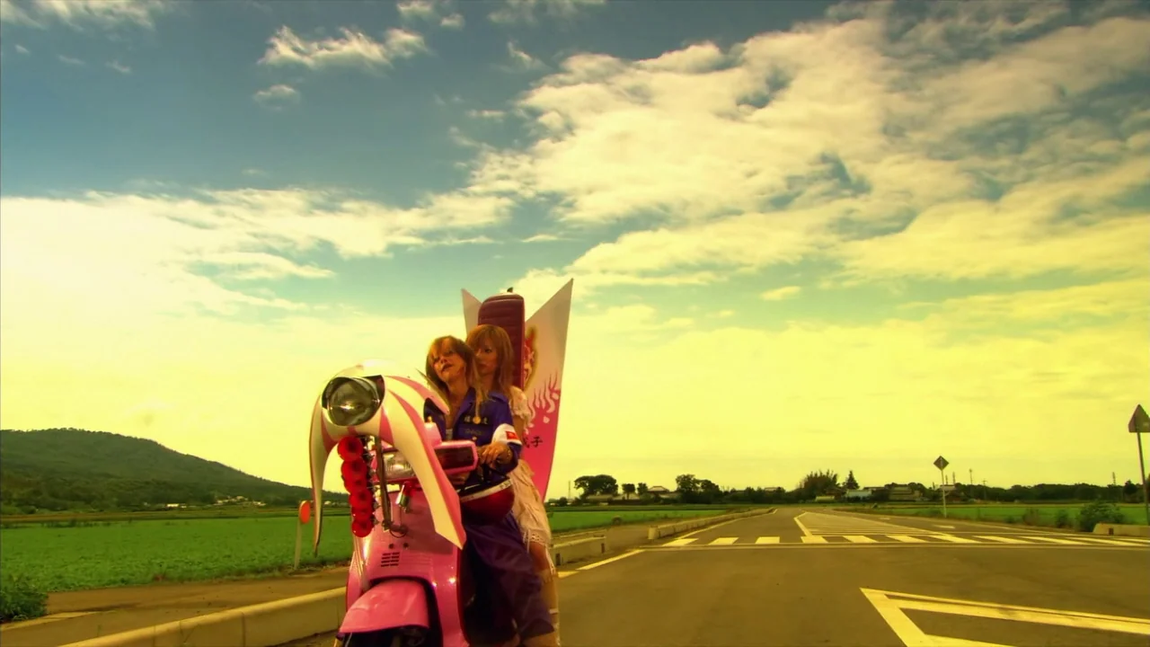 Kamikaze Girls (2004) movie still: medium — A rural road stretches through green fields under a bright, cloud-filled sky. Two women sit on a…