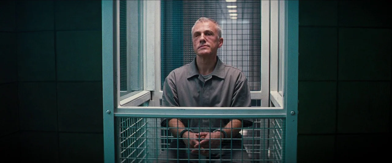 No Time To Die (2021) movie still: medium — A man sits inside a small steel and wire mesh enclosure. He faces the camera with his wrists restrained…