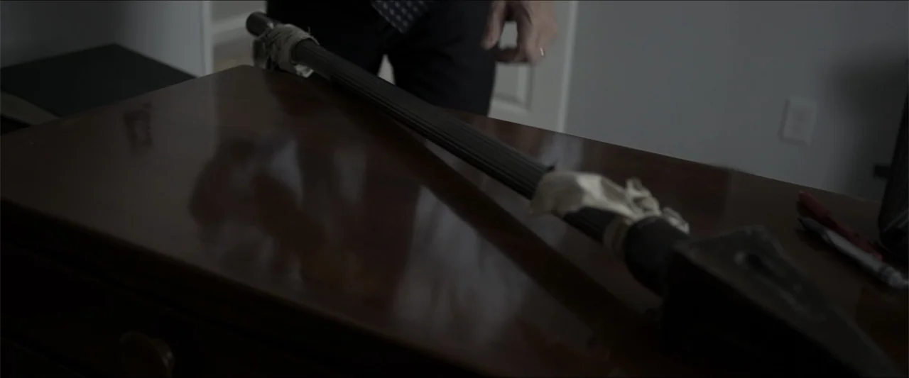 Heavenly Deposit (2019) movie still: closeup — A polished wooden surface holds a metal tool wrapped in white cloth tape. The blurred figure of a…