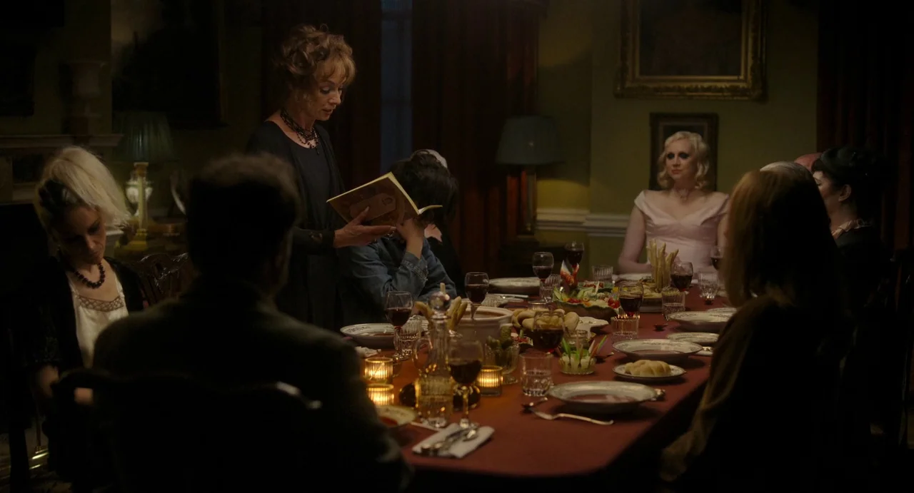 Flux Gourmet (2022) movie still: medium — A dimly lit room features a long dining table set with food and glassware. A woman stands in the center…