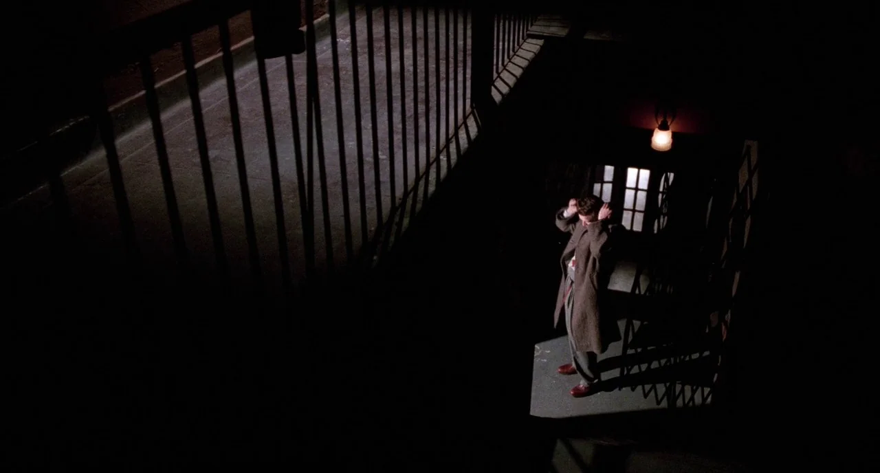Angel Heart (1987) movie still: wide — A dark, industrial interior stairwell with iron railings features a single overhead light. A man in a long…