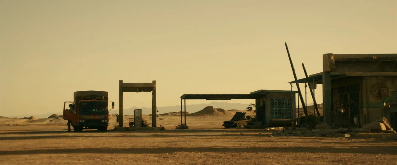 War Dogs (2016) movie still: wide — An abandoned gas station sits in the middle of a vast, flat, arid desert. A parked orange truck with an open…