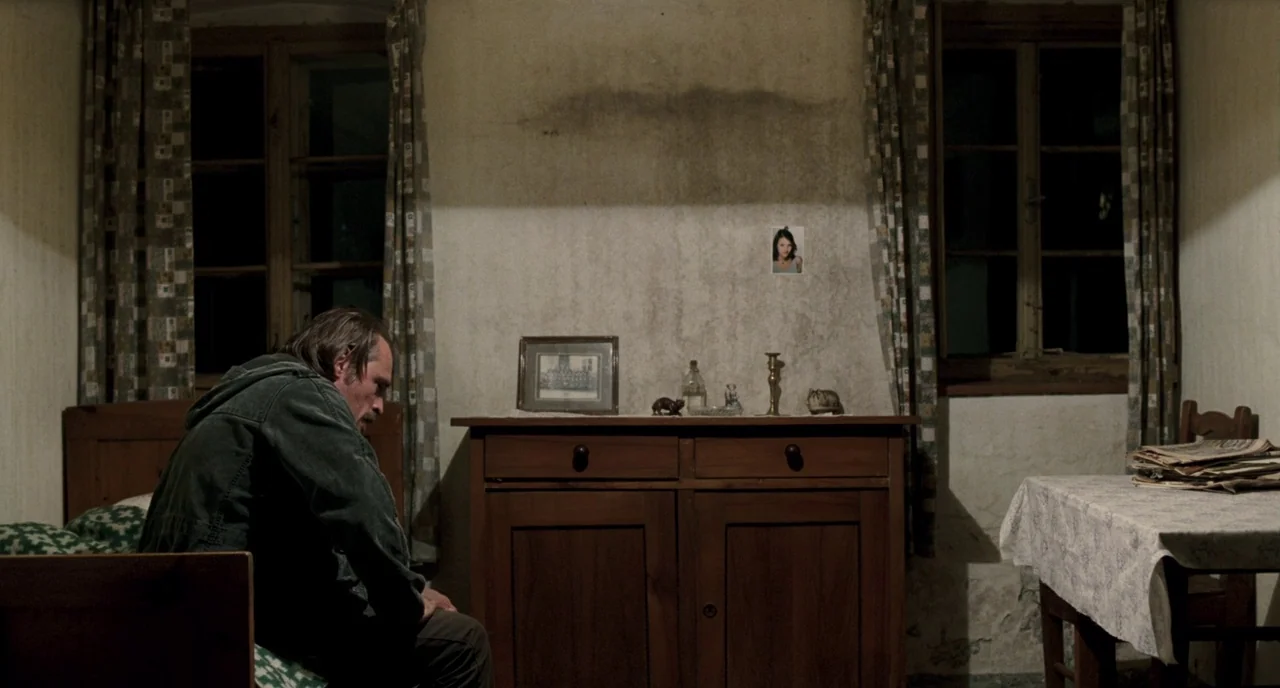 Revanche (2008) movie still: medium — A rustic, sparsely furnished room features weathered walls and old wooden furniture. A man sits alone on the…