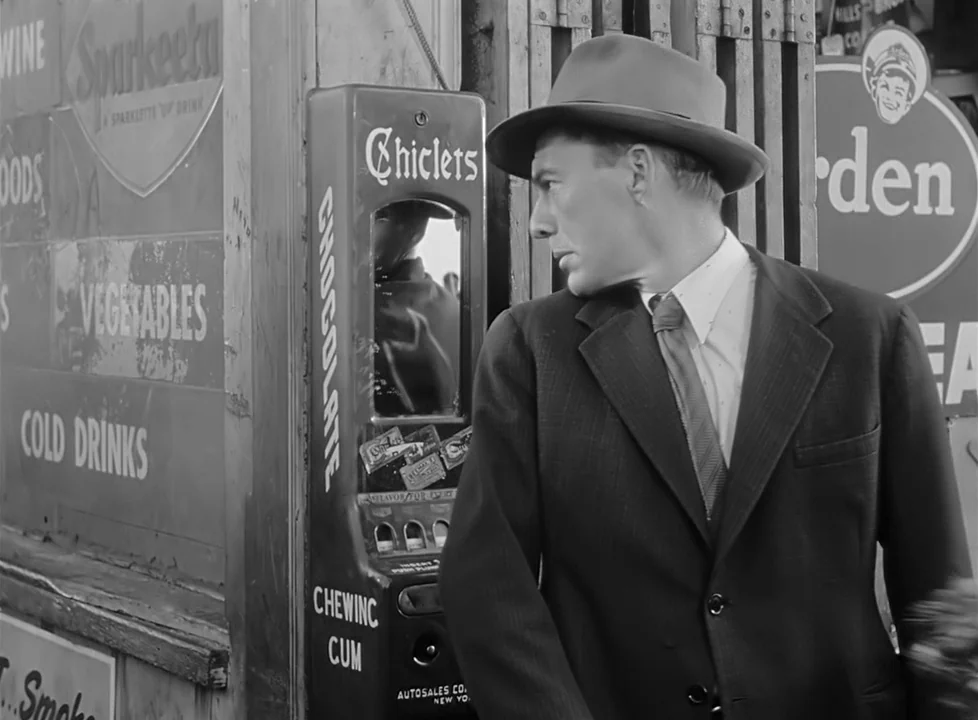 M (1951) (1951) movie still: medium — A man in a suit stands next to a Chiclets gum vending machine outdoors. Various advertisements for Sparkleeta…