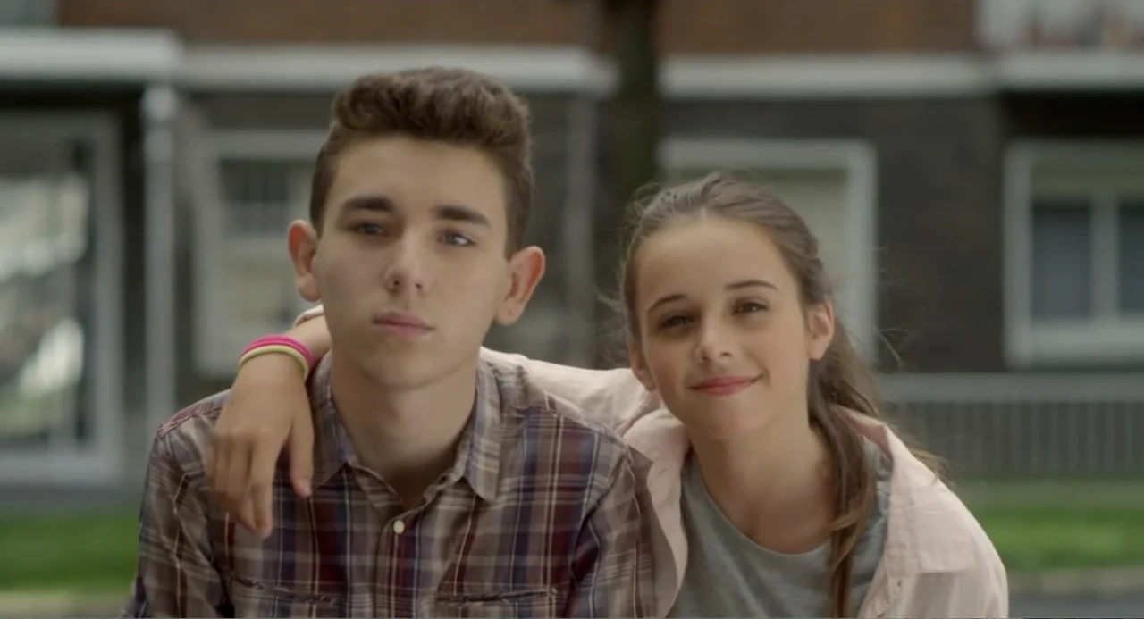 Newton's Third Law (2017) movie still: medium — An outdoor neighborhood street setting with houses blurred in the background. A teenage boy and girl…
