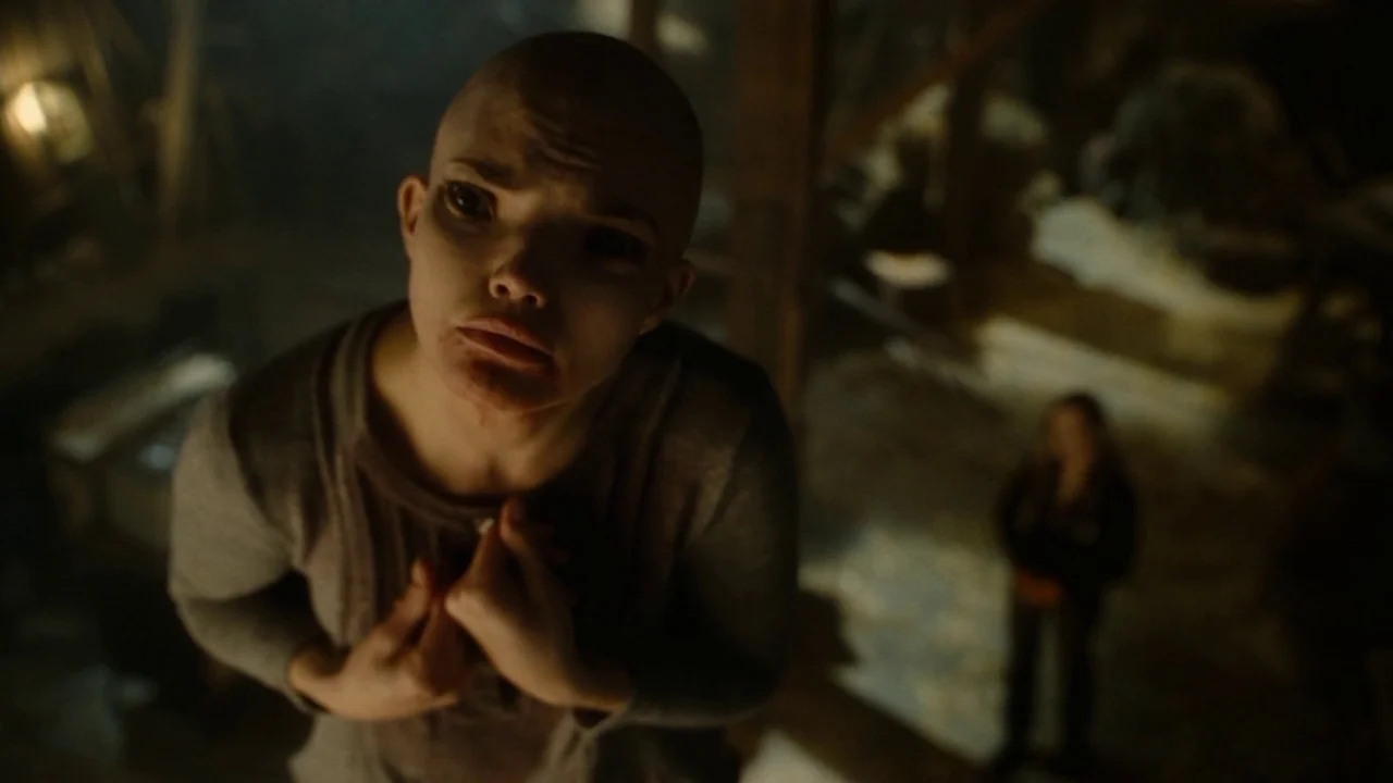 Splice (2009) movie still: medium — A dimly lit, dusty attic space features wooden rafters and structural beams overhead. A bald woman in a grey…