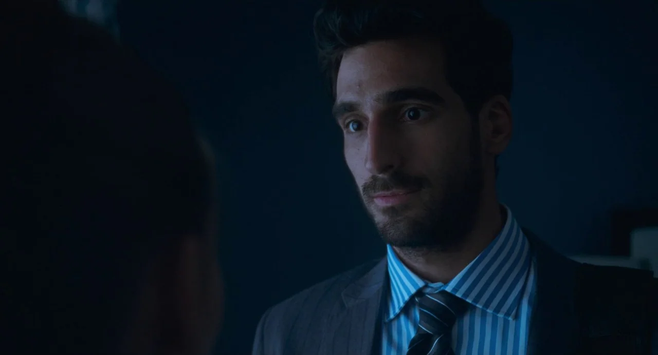 Matthias & Maxime (2019) movie still: closeup — A dark, dimly lit indoor space with low ambient lighting. A man in a suit looks toward an unseen…