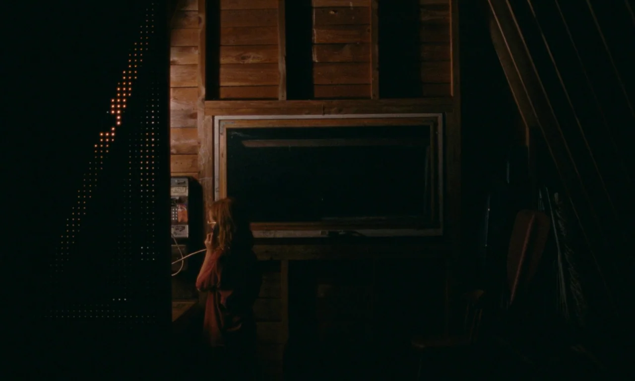 Janet Planet (2023) movie still: medium — A dark, wood-paneled attic space features a solitary telephone mounted on a wall. A woman in silhouette…