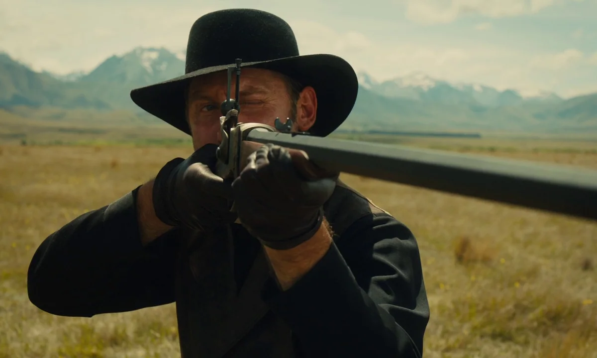 Slow West (2015) movie still: medium — A man stands in a vast, arid open field with distant mountains in the background. He holds a rifle level,…