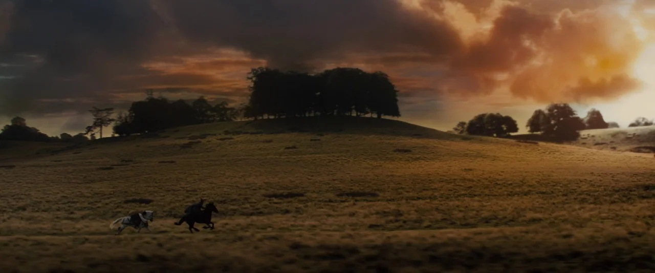 Maleficent (2014) movie still: wide — A vast, rolling expanse of dry golden grass sits under a dark, dramatic sky. Two distant figures on horseback…