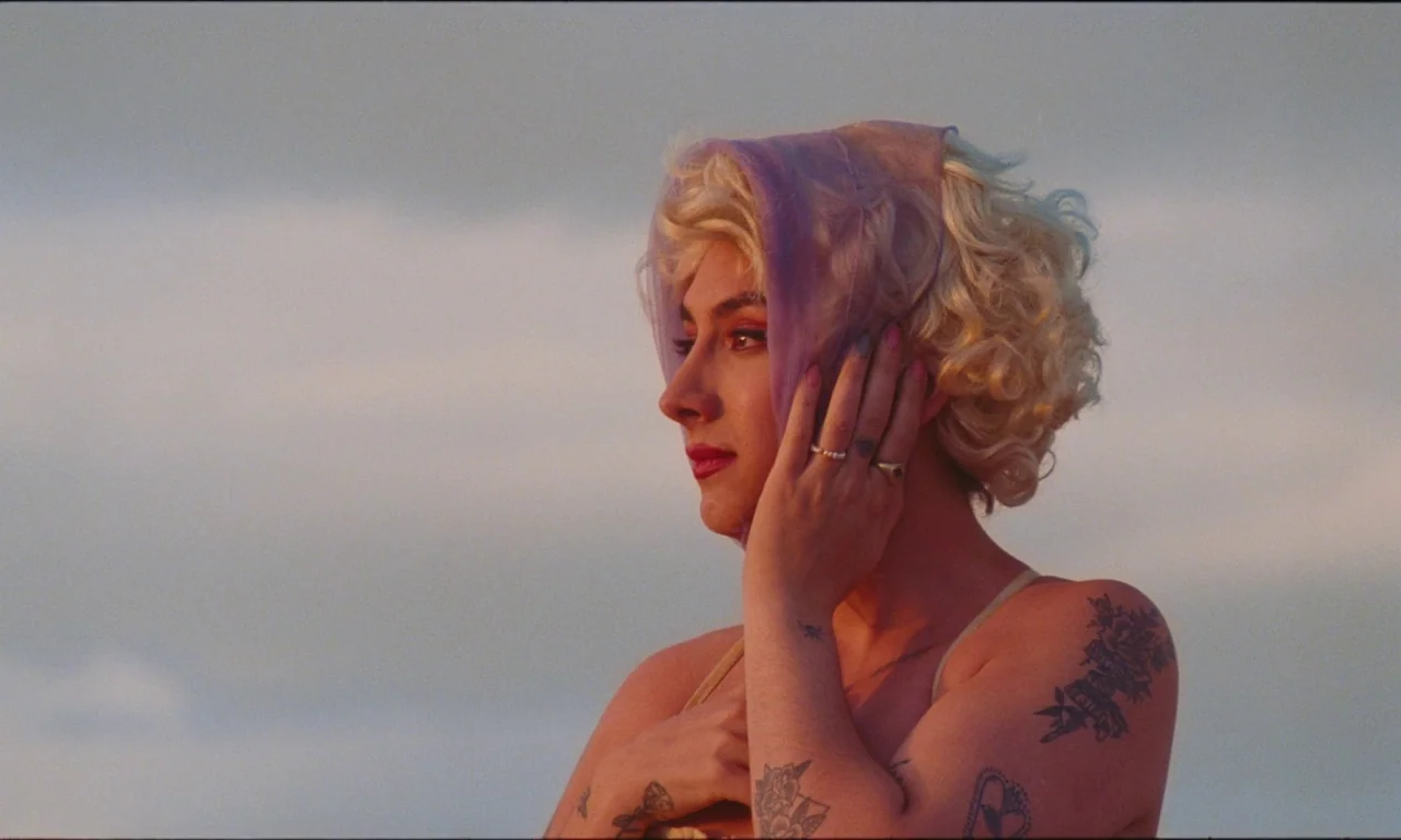 National Anthem (2023) movie still: closeup — A person stands outside during the golden hour against a hazy, clouded sky. The subject holds a…