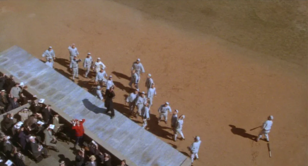 Eight Men Out (1988) movie still: wide — A high-angle view shows a baseball dugout at the edge of a dirt field. A single player walks from the…