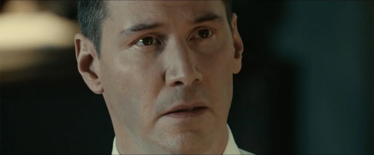 Exposed (2016) movie still: closeup — An interior space with a blurred out-of-focus background in muted colors. A middle-aged man in a white dress…