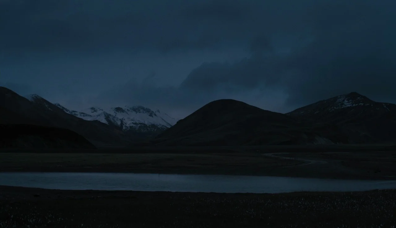 Land Ho! (2014) movie still: wide — A vast landscape features jagged snow-capped mountains overlooking a calm, dark pool of water. Dark, dense…
