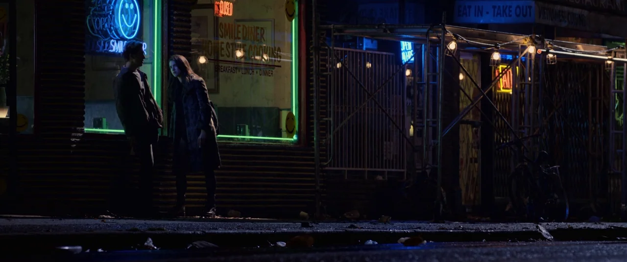 Death Note (2017) movie still: wide — A night city sidewalk with a diner window illuminated by neon signage. A young man and woman stand talking in…