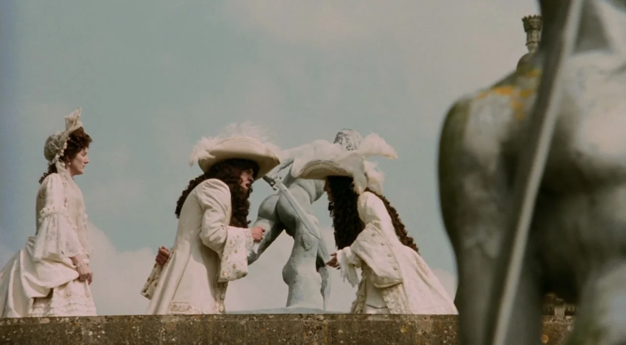 Orlando (1992) movie still: fullbody — A stone terrace sits beneath a pale overcast sky with a weathered statue. Two men in historical costumes…