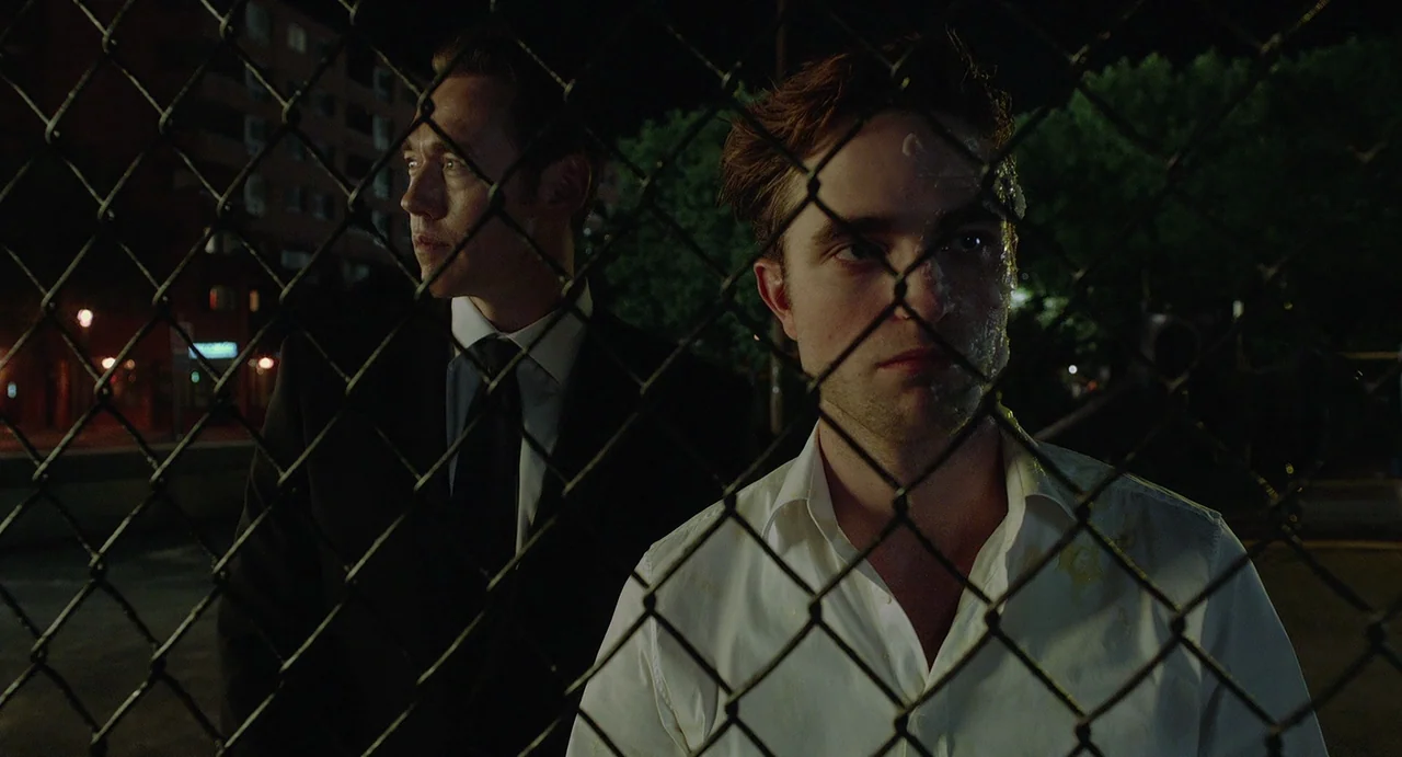 Cosmopolis (2012) movie still: medium — Two men stand behind a chain-link fence at night near some urban buildings. They are positioned closely…